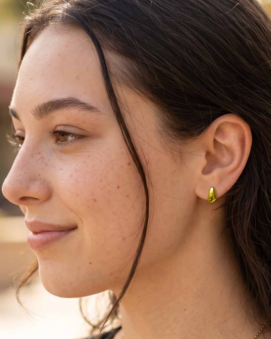 A close-up side profile of a woman wearing a small gold crescent-shaped stud earring. Teardrop Stud Earrings - Gold.