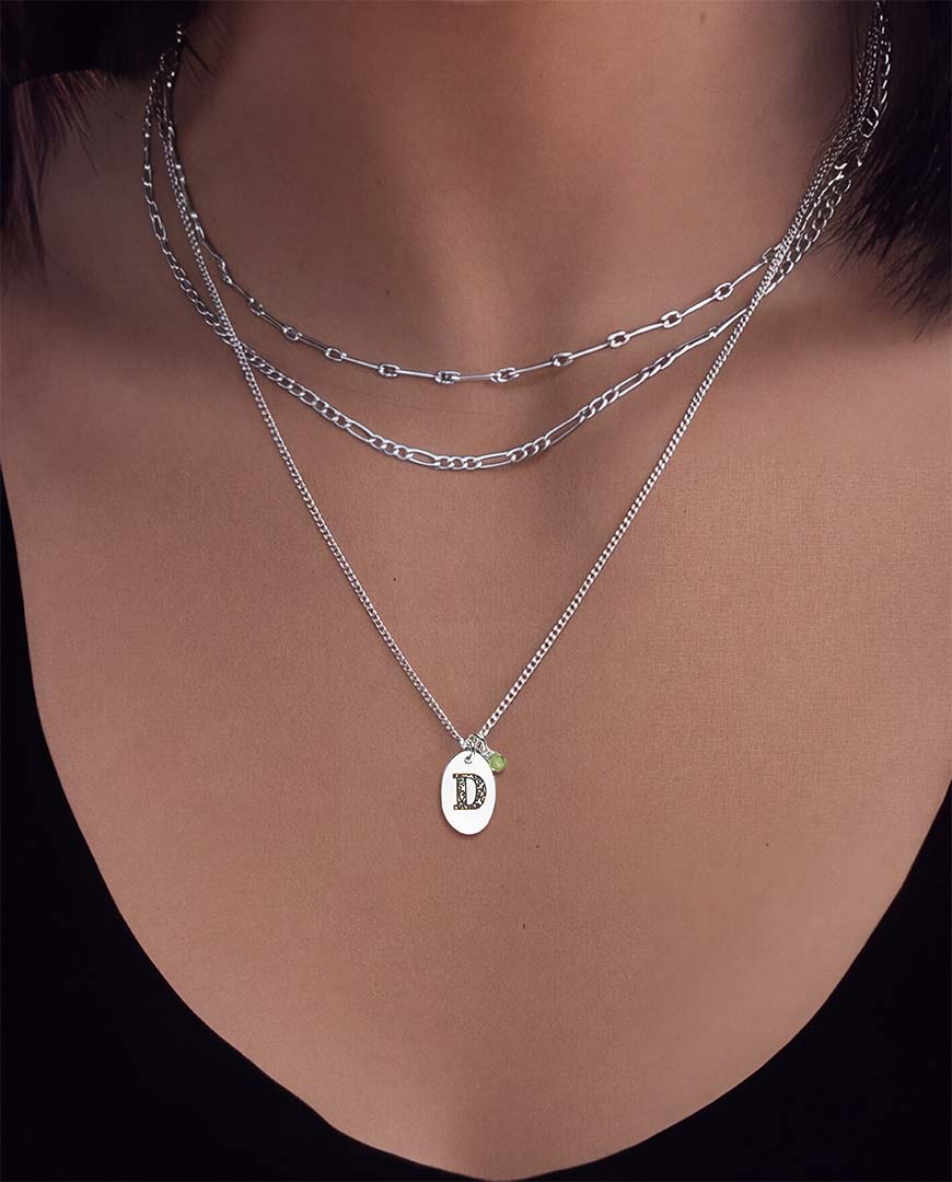 A close-up of a model's neck and chest wearing three layered silver necklaces, including a short chain, a longer curb chain, and a delicate chain with an oval pendant engraved with the letter 'D'. The Loveletter necklace in silver.