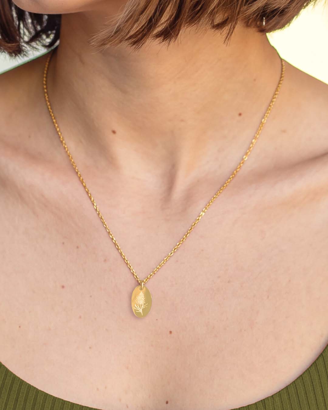 A close-up of a person's neck and upper chest wearing a delicate gold chain necklace with a small oval pendant with  grevillea flower engraved on it