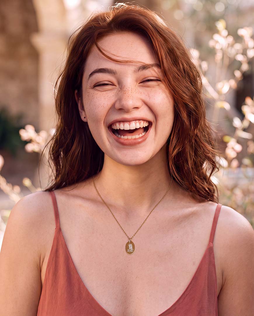 A woman with wavy auburn hair is shown from the chest up, laughing joyfully outdoors. She is wearing a delicate gold pendant necklace the Bottlebrush necklace from the Australian Native Birthflower collection by sixD
